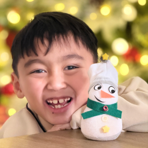 Young boy smiling next to the sock snowperson he made in front of a blurred christmas tree with lights